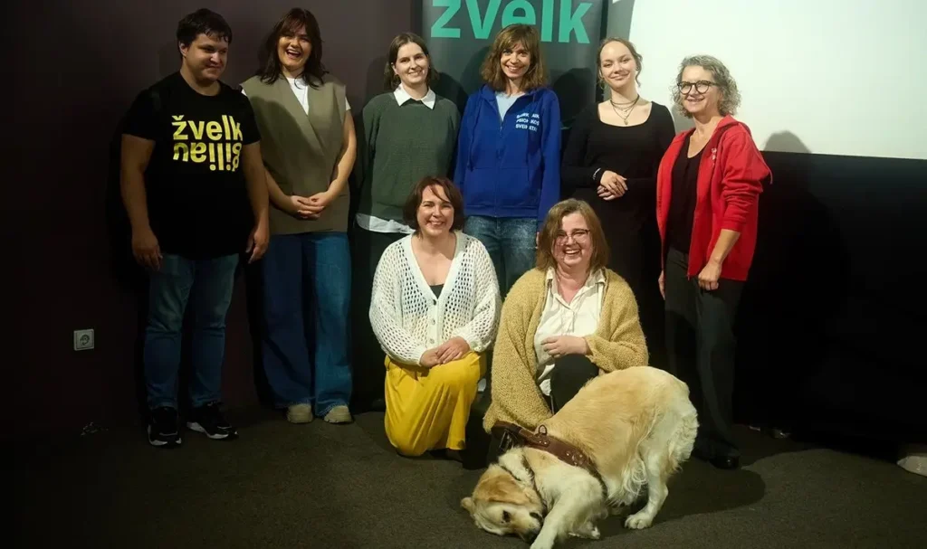 Aštuoni žmonės, vieni stovi, kiti klūpo, šypsosi bendrai nuotraukai pritemdytame kambaryje. Priekiniame plane ant grindų ilsisi šuo asistentas.