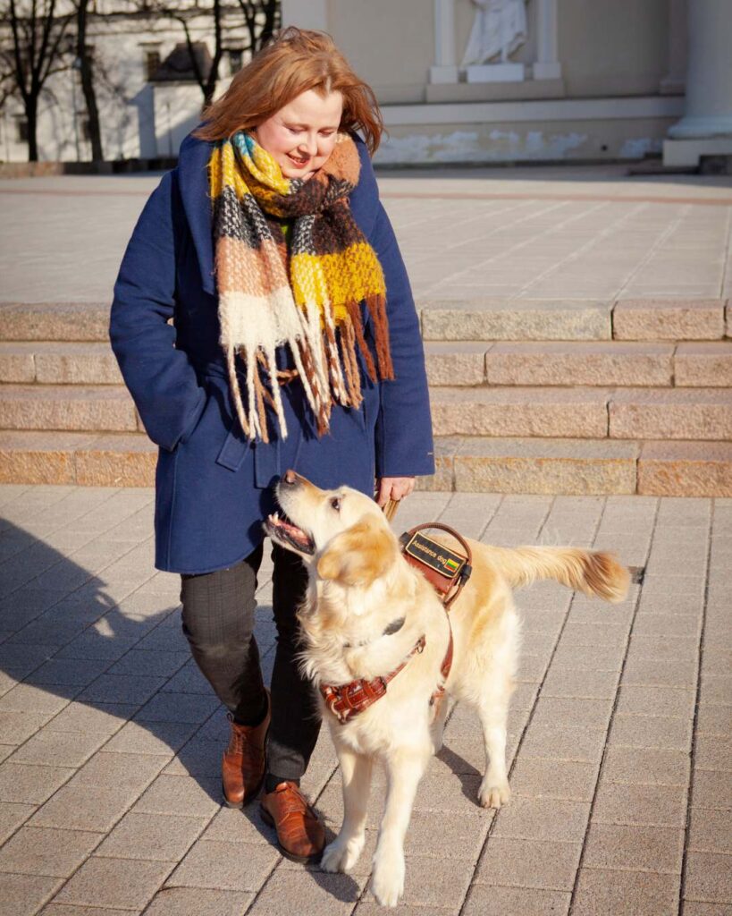 Moteris su mėlynu paltu ir spalvingu šaliku šypsosi auksaspalviam retriveriui vedliui saulėtą dieną. Jie stovi ant laiptų, perteikdami draugiškumą. Šuo vilki odines šuns asistento petnešas.